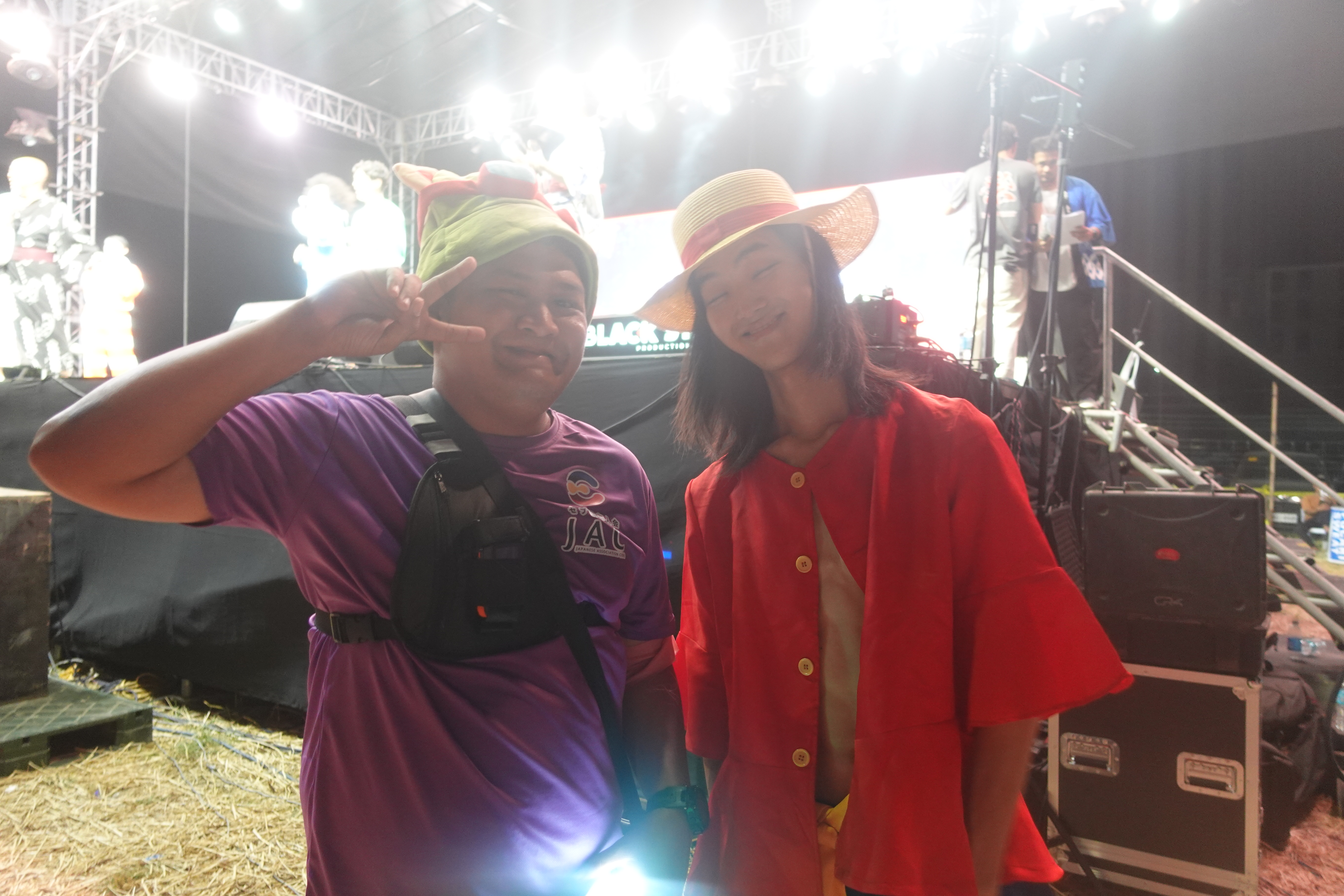 Two people posing for photo backstage, one wearing straw hat and red outfit.