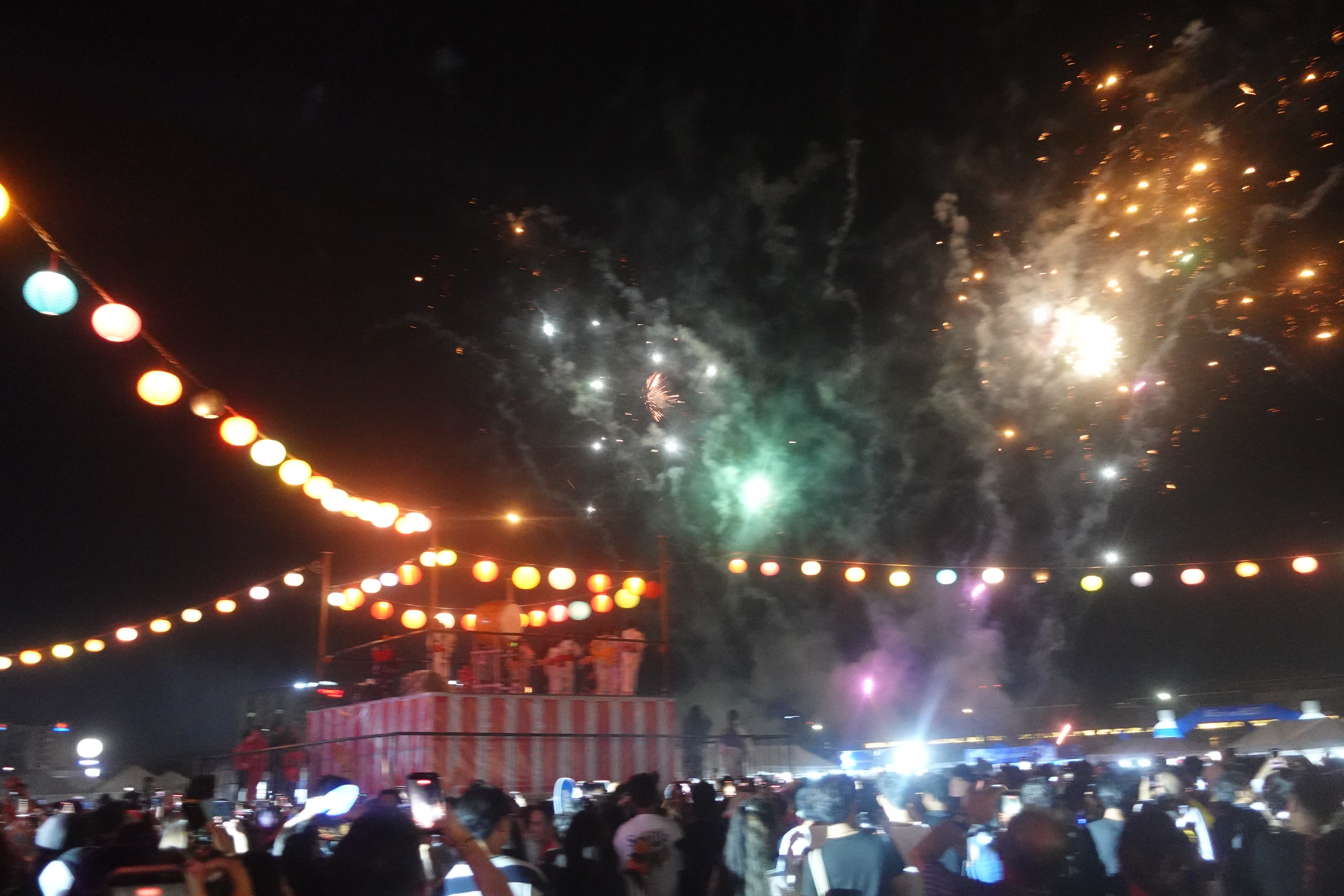 Fireworks over festival at night, lanterns hanging above crowd.