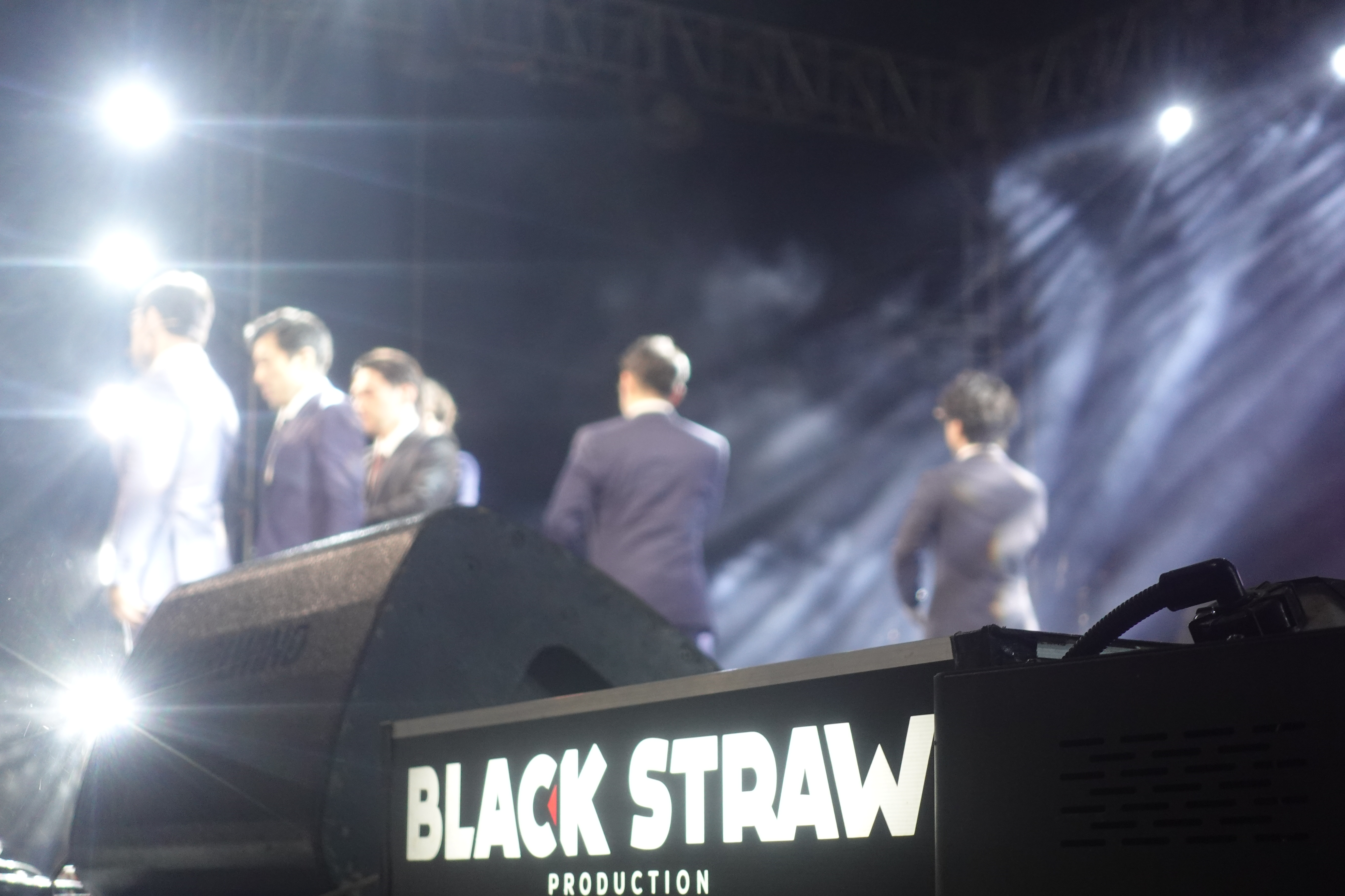 Backstage view of performers walking near stage, “BLACK STRAW” signage visible.