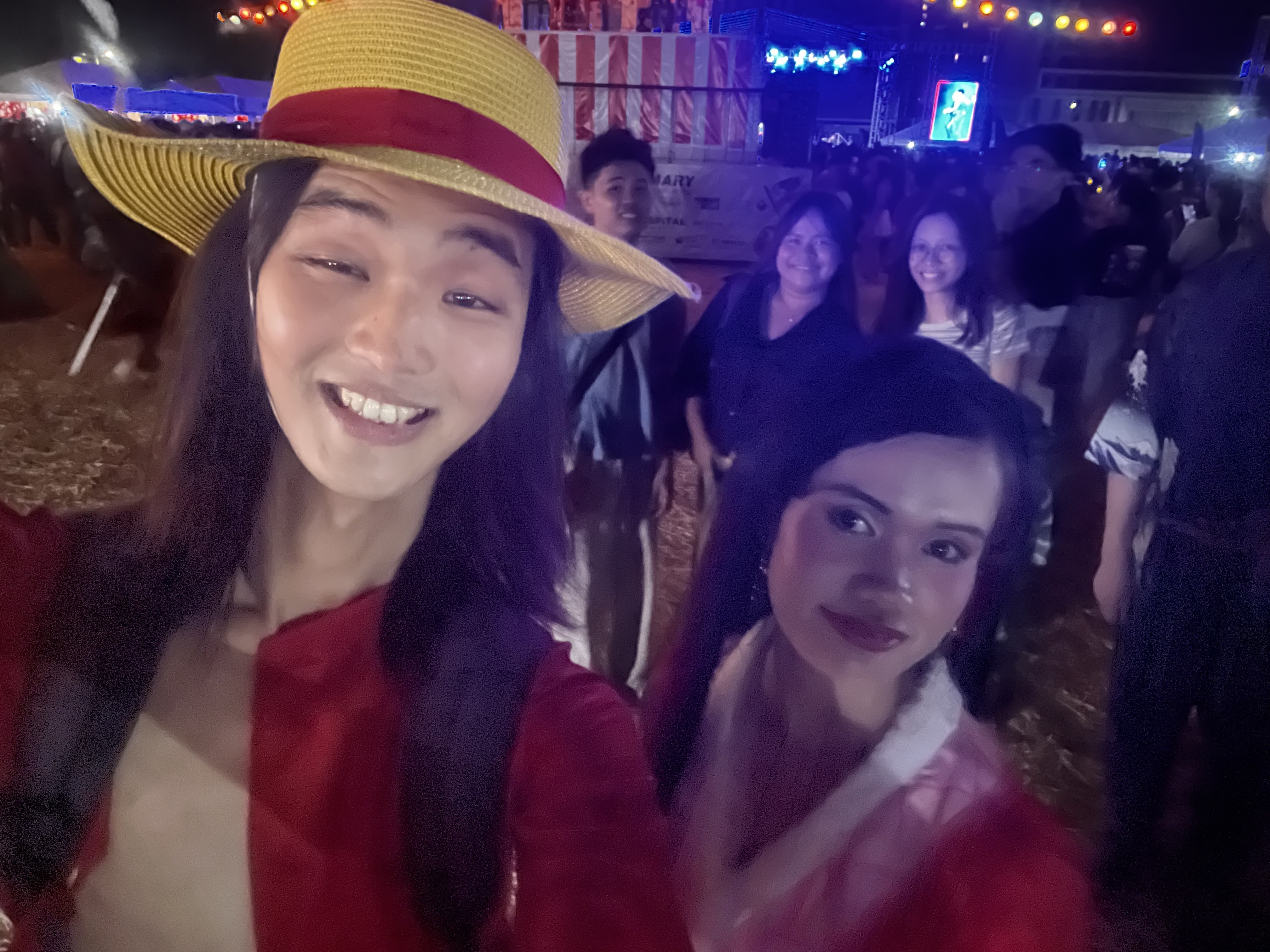 Person in straw hat smiling for selfie with a friend behind, festival crowd around them.