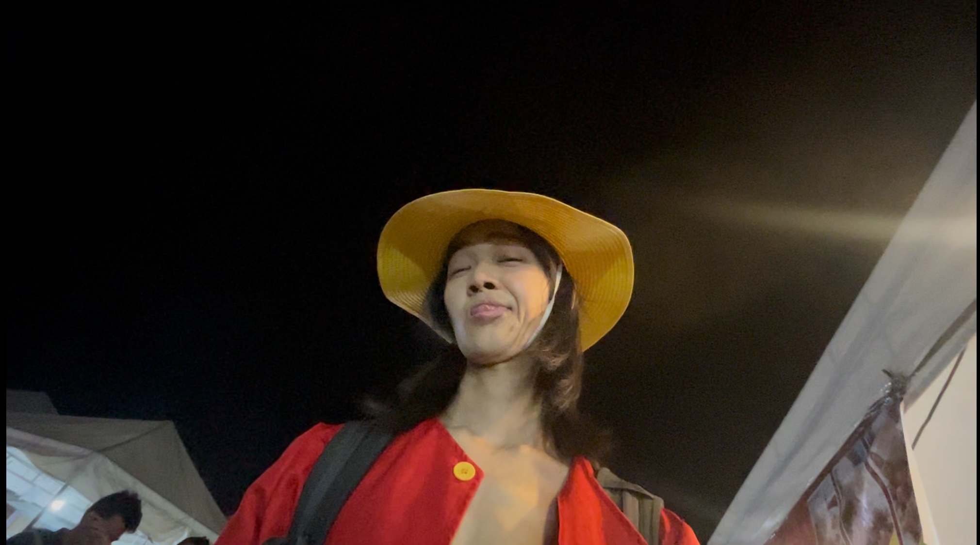 Person in a straw hat and red outfit taking a selfie, smiling with festival lights behind.