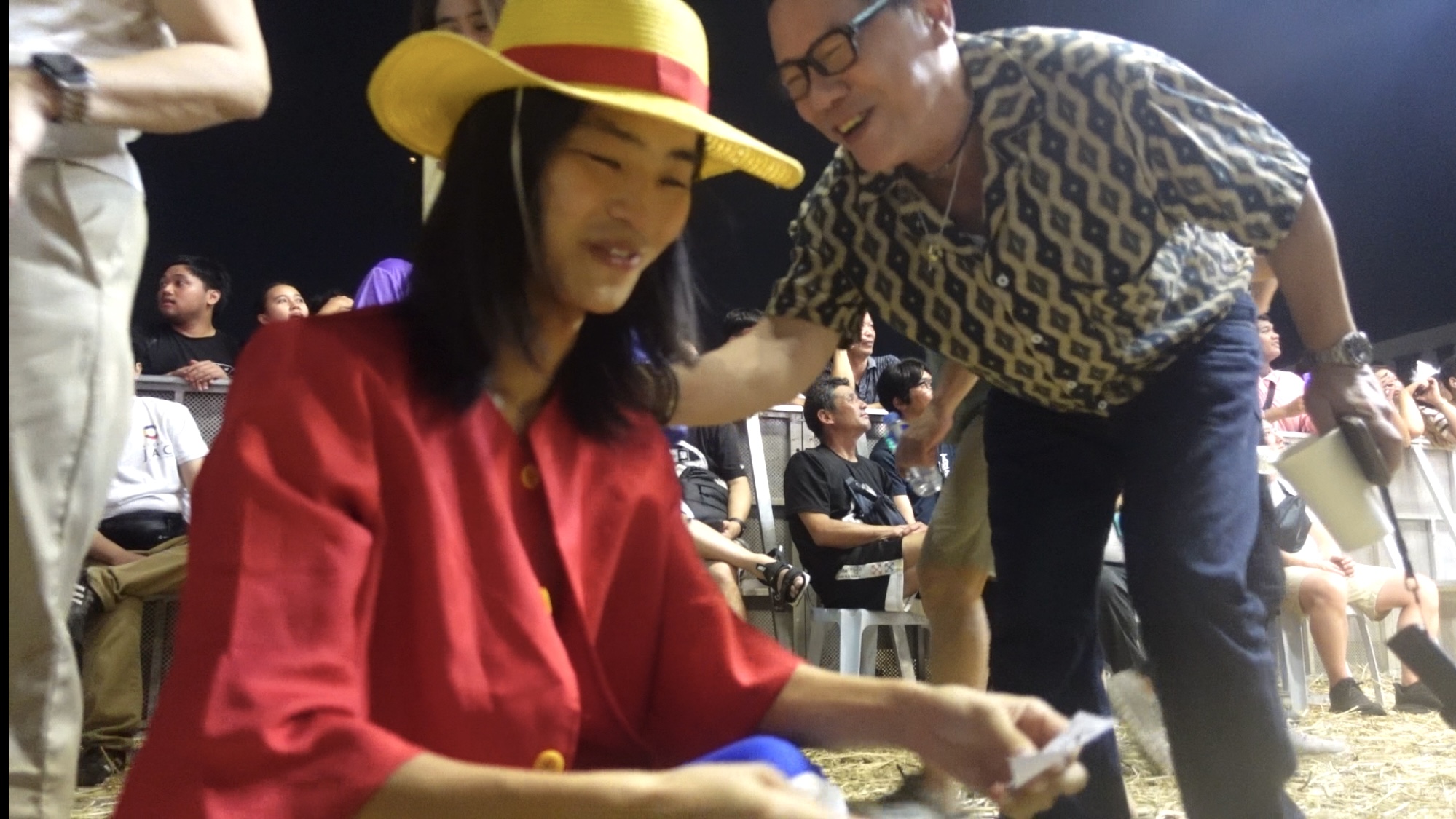 Person in straw hat and red outfit sitting while a man leans over and smiles.