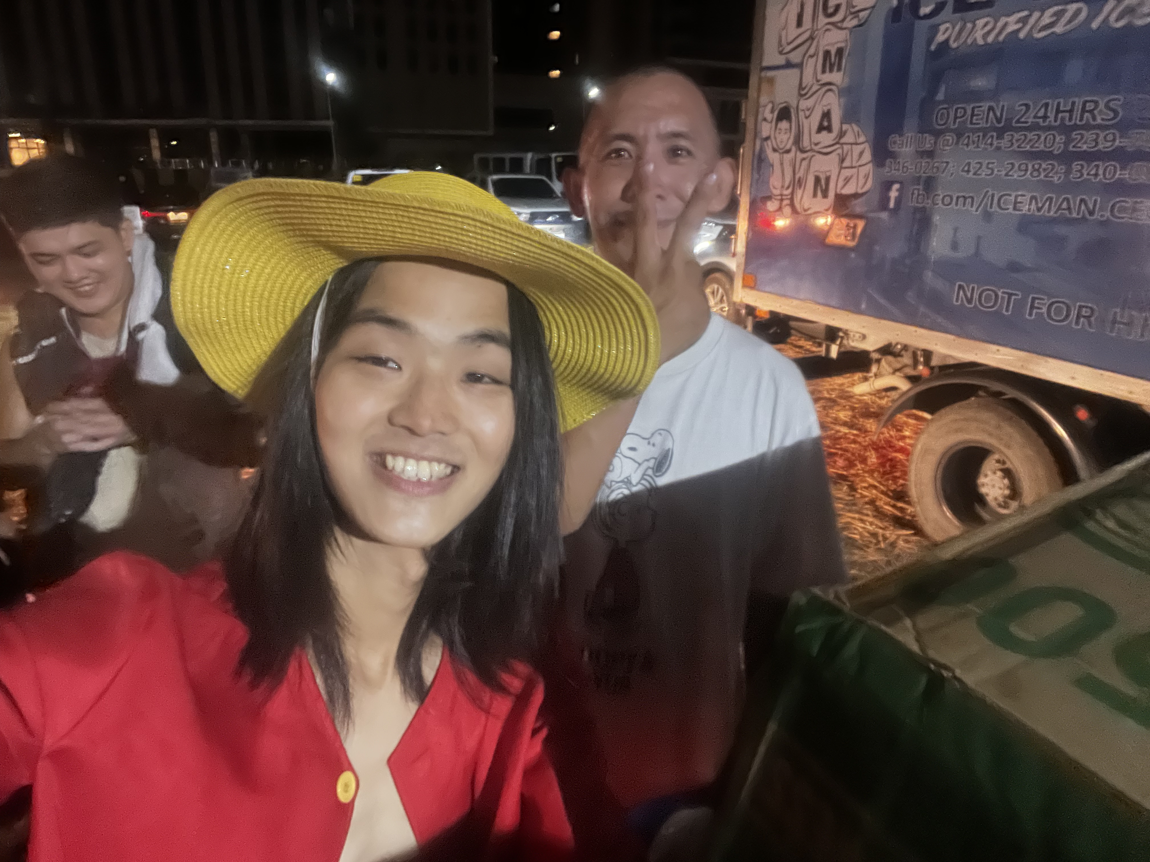 Person in straw hat smiling for selfie with two people behind, night festival scene.