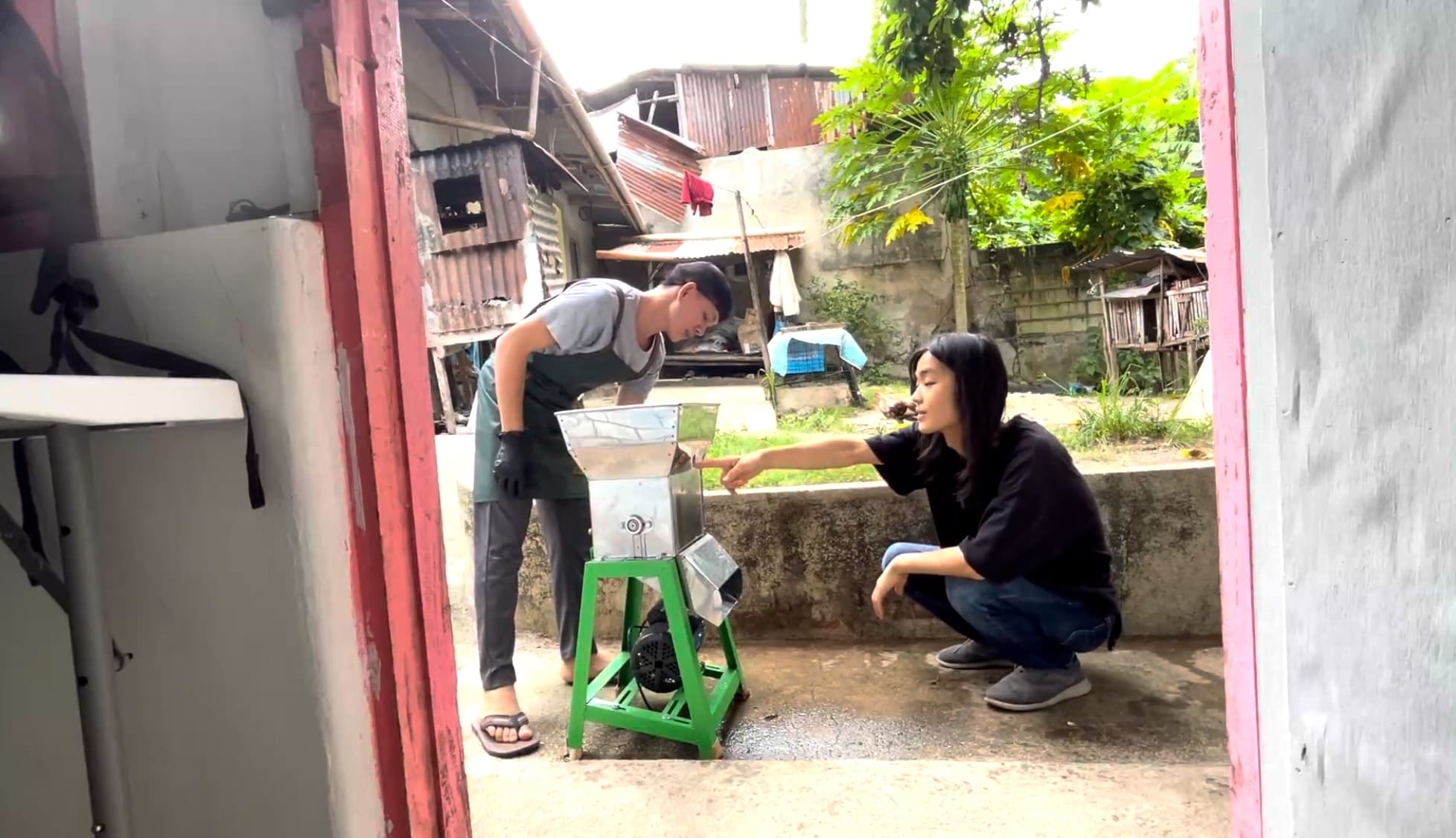 Two men beside a cassava grating machine outdoors, with one pointing at the machine