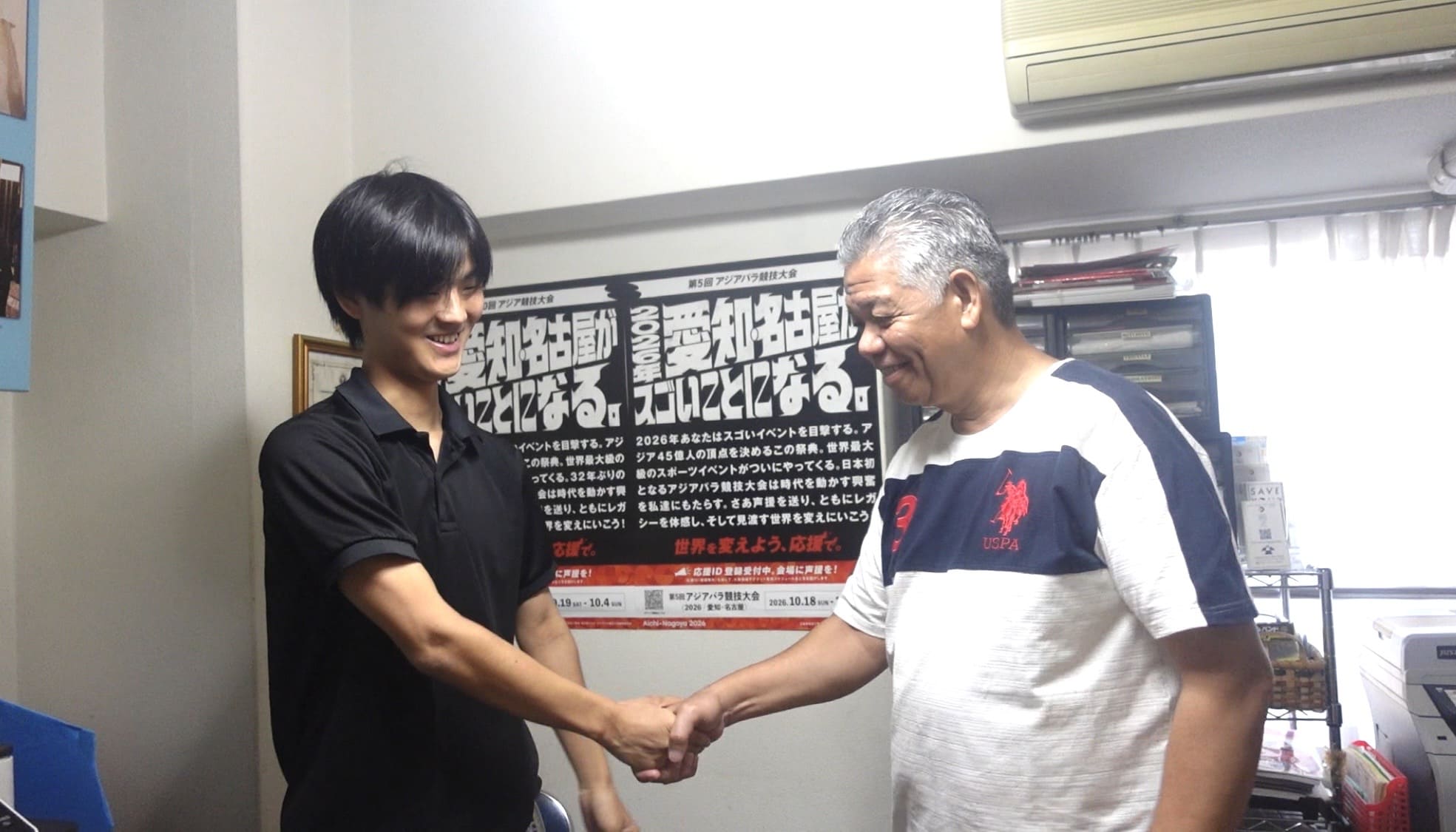 Two men smiling and shaking hands inside a community office in Nagoya
